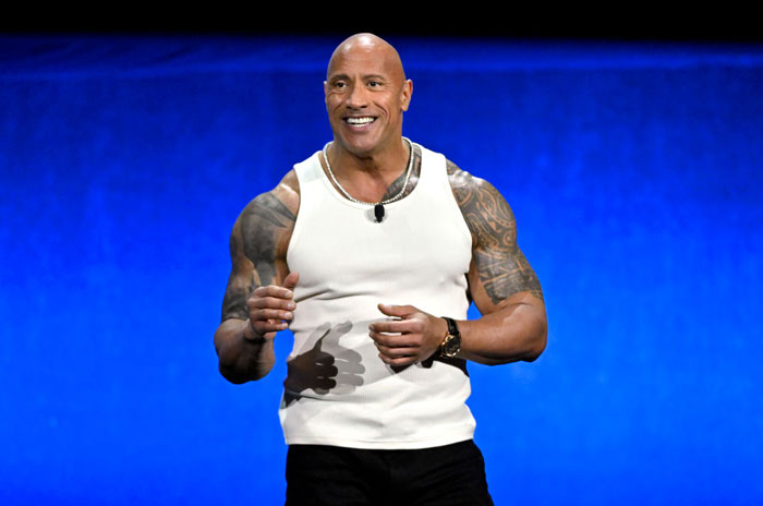 Dwayne "The Rock" Johnson, wearing a white tank top, smiles while speaking on stage, showing his muscular arms and tattoos against a blue background.