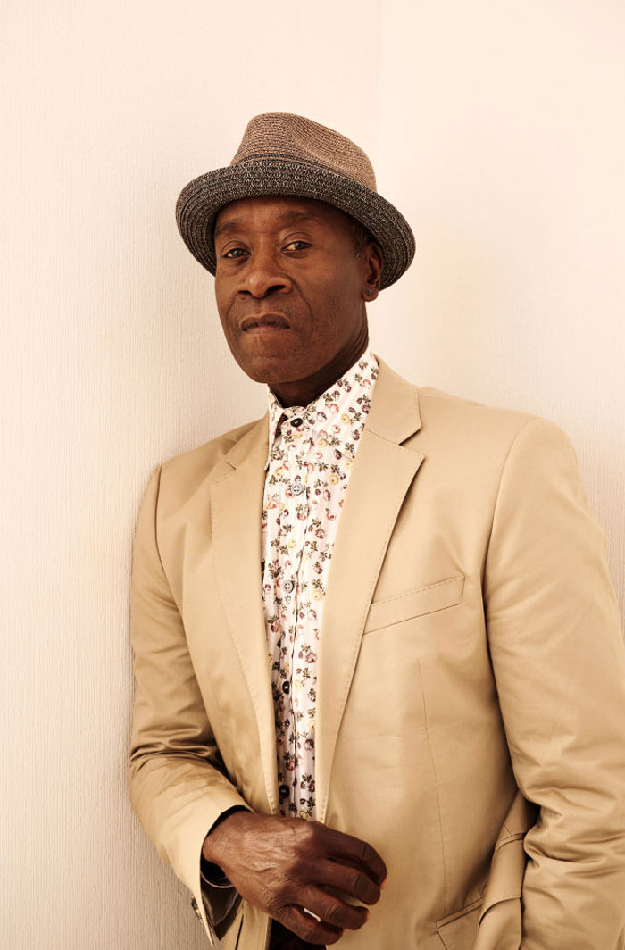  A B list celebrity, Don Cheadle, is posing against a light-colored wall, wearing a beige suit, a floral shirt, and a stylish hat, with a serious expression.