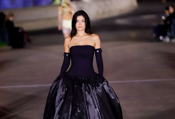 Kylie Jenner walks down the runway in a black, strapless gown with matching long gloves during a fashion show, with a focused expression.