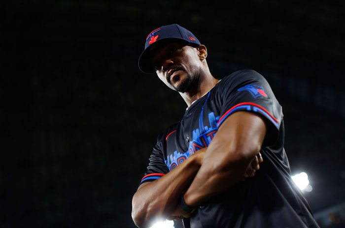  A B list celebrity, Anthony Mackie, is standing with his arms crossed, wearing a Toronto-themed baseball uniform and cap, looking down under stadium lights.