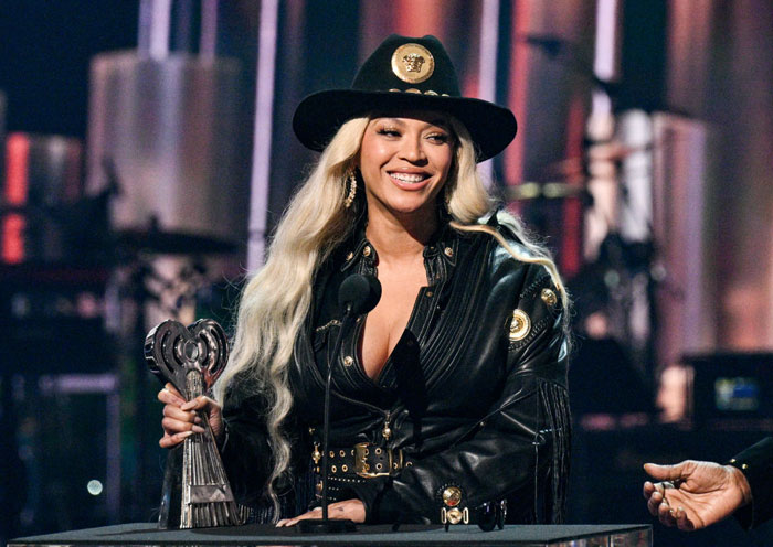 Beyoncé, wearing a black leather outfit and wide-brimmed hat, smiles while accepting an award on stage, holding a trophy in her hand.