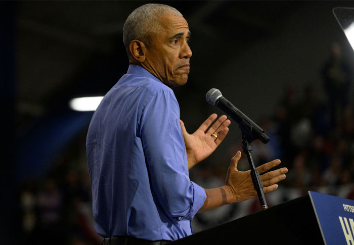 Barack Obama, wearing a light blue shirt, gestures with his hands while speaking at a podium during an event, with a thoughtful expression.