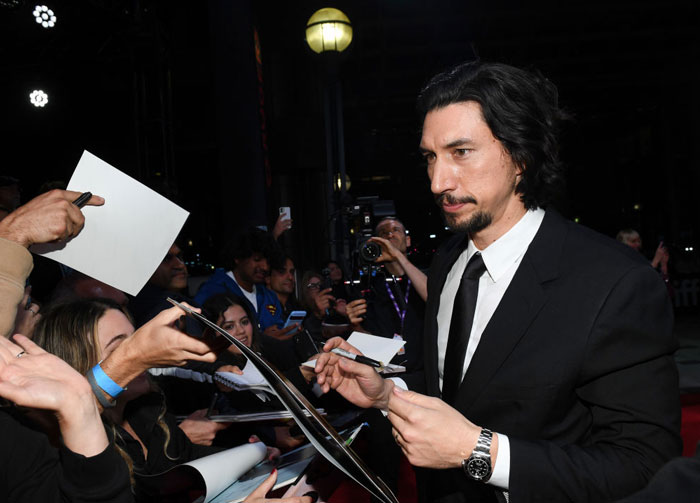  A B list celebrity, Adam Driver, is signing autographs for fans while wearing a black suit and tie at an outdoor evening event, surrounded by cameras and fans.