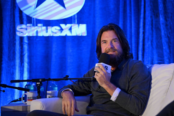  A B list celebrity, Jason Bateman, is seated in a SiriusXM interview, holding a microphone and speaking, with a blue-lit backdrop and branded signage behind him.