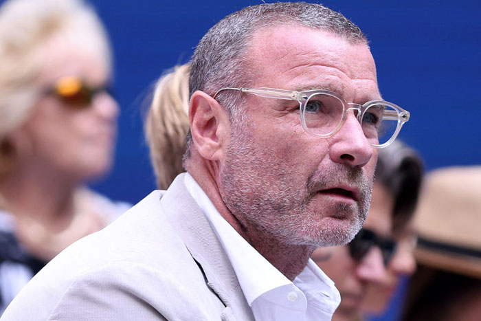 A B list celebrity, Liev Schreiber, is pictured wearing clear-framed glasses and a light-colored suit while attending an outdoor event, with people in the background.