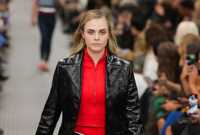  A B list celebrity, Cara Delevingne, is walking the runway in a red zip-up top and a black shiny jacket during a fashion show, with a crowd in the background.