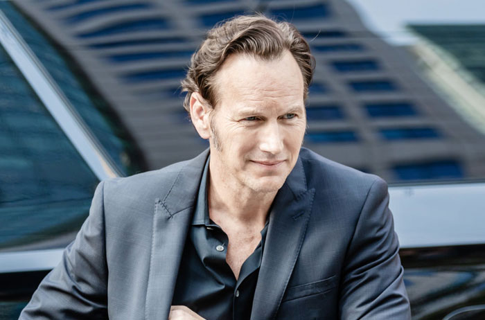  A B list celebrity, Patrick Wilson, who is wearing a dark suit and black shirt while leaning against a car with a city building reflecting in the background.