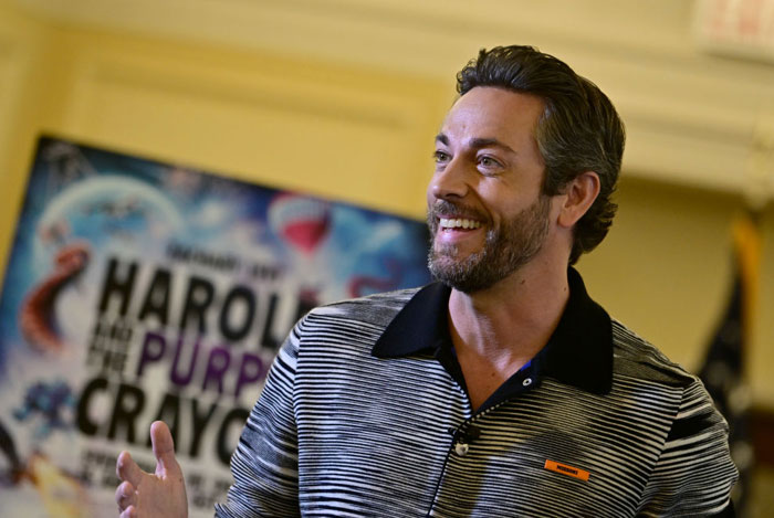  A B list celebrity, Zachary Levi, is smiling and gesturing during a public event, wearing a striped polo shirt. A colorful "Harold and the Purple Crayon" poster is in the background.