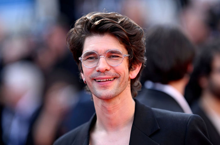 A B list celebrity, Ben Whishaw, is smiling while wearing clear-framed glasses and a black blazer at an outdoor red carpet event, with a blurred crowd in the background.