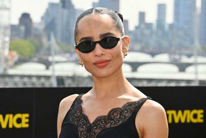  A B list celebrity, Zoë Kravitz, is posing outdoors wearing black sunglasses and a black lace-trimmed top, with a cityscape in the background.