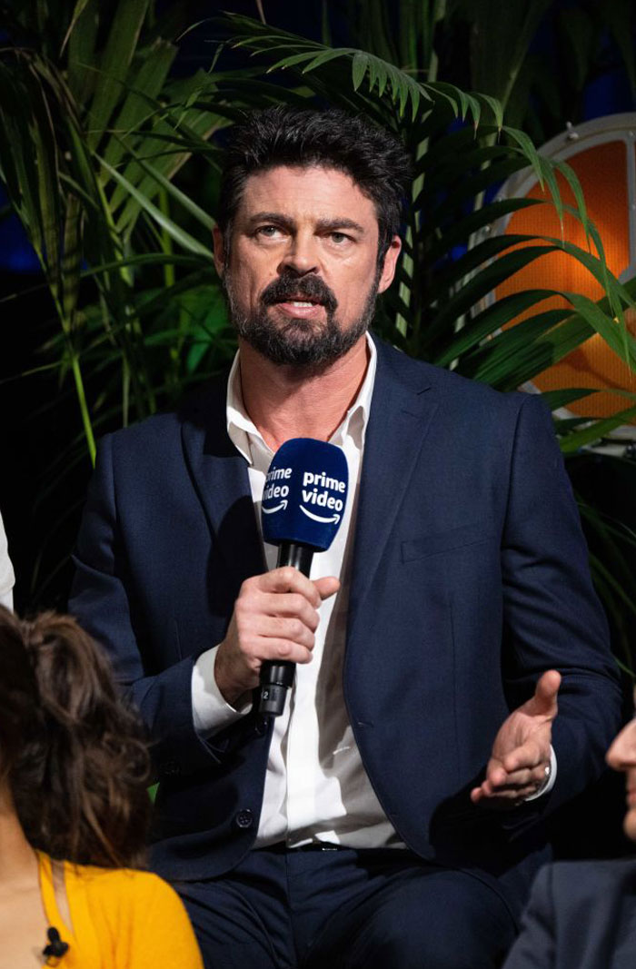  A B list celebrity, Karl Urban, is speaking into a Prime Video microphone while wearing a navy blue suit and white shirt, with greenery in the background.