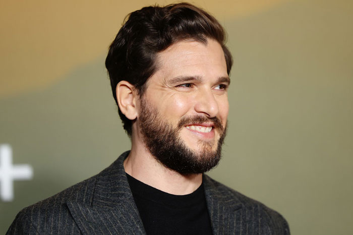  A B list celebrity, Kit Harington, is smiling while wearing a dark pinstripe blazer and black shirt at an indoor event, with a blurred backdrop behind him.