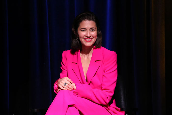  A B list celebrity, Mary Elizabeth Winstead, is seated and smiling while wearing a bright pink suit during an event, with a dark curtain backdrop behind her.