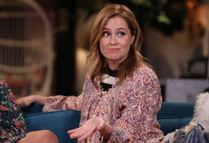  A B list celebrity, Jenna Fischer, is seated on a couch, gesturing with her hand in a playful manner while wearing a floral blouse during an indoor event.