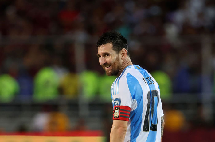 Lionel Messi, wearing an Argentina jersey with the number 10 and a captain's armband, smiles during a football match, looking over his shoulder.