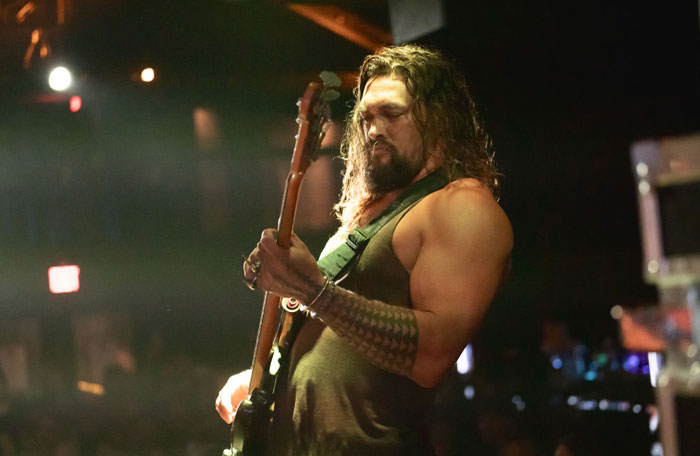  A B list celebrity, Jason Momoa, who is performing on stage, playing a bass guitar while wearing a sleeveless shirt and showcasing his muscular arms and tattoo.