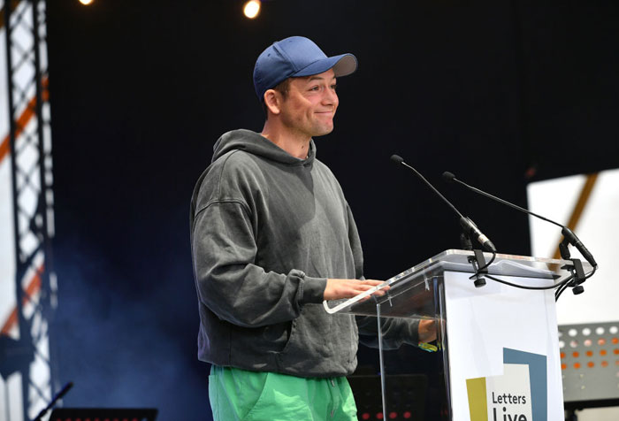  A B list celebrity, Taron Egerton, is speaking at a podium during a "Letters Live" event, wearing a gray hoodie, green pants, and a blue baseball cap on stage.