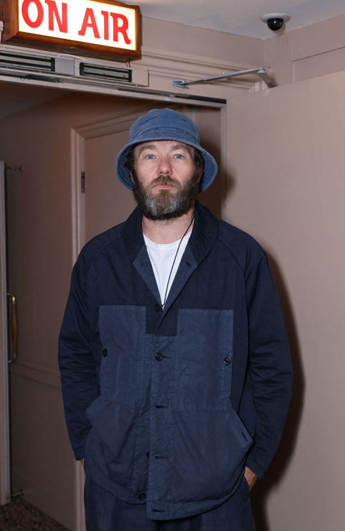  A B list celebrity, Joel Edgerton, is standing under an "On Air" sign, wearing a blue bucket hat and matching jacket, with a neutral expression indoors.