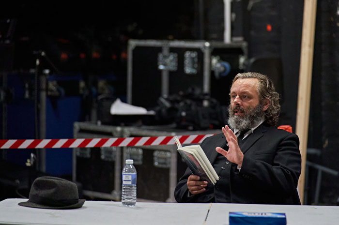  A B list celebrity, Michael Sheen, is seated at a table backstage, holding a book and gesturing with his hand while wearing a black suit. A bottle of water and a hat are placed on the table nearby.