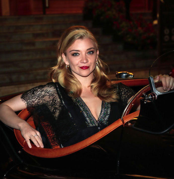  A B list celebrity, Natalie Dormer, is sitting in a vintage car, wearing a black lace dress with a plunging neckline, and smiling for the camera at an outdoor evening event.