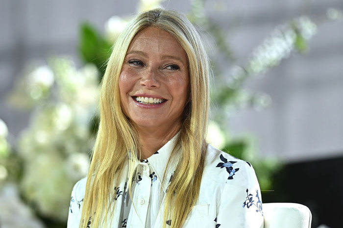  A B list celebrity, Gwyneth Paltrow, is smiling while seated at an event, wearing a white floral blouse with her blonde hair down, with a floral backdrop behind her.