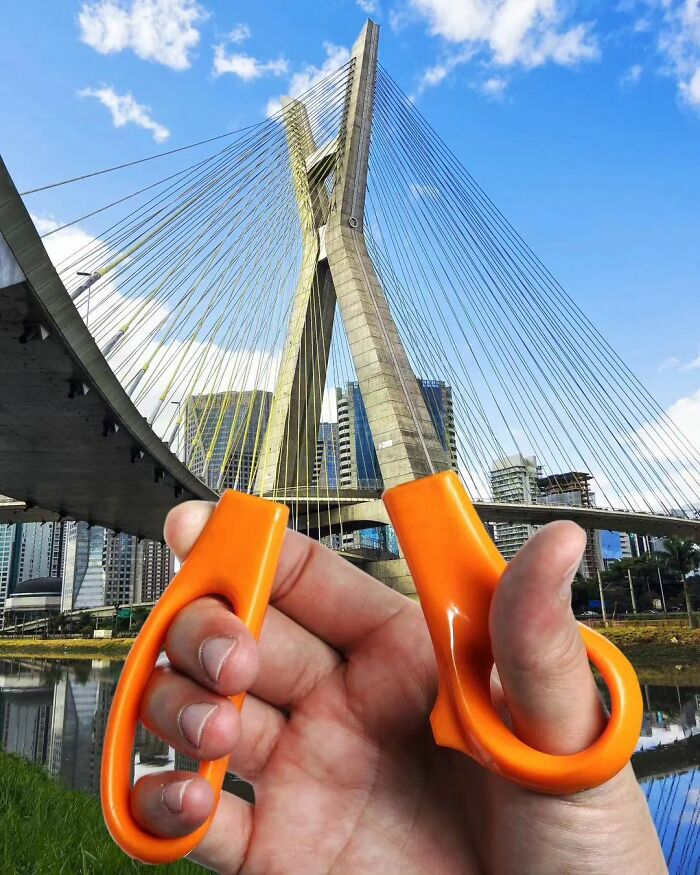 A hand holding scissors aligned with a bridge for a surreal effect, highlighting surrealism.