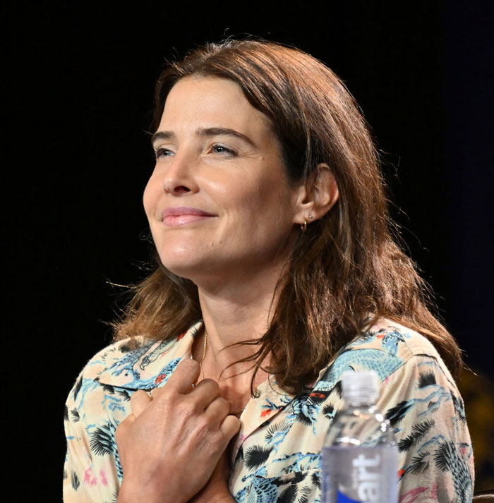 A B list celebrity, Cobie Smulders, is seated at an event, smiling softly while wearing a floral shirt, with her hands gently clasped at her chest and a water bottle nearby.