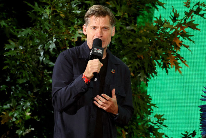  A B list celebrity, Nikolaj Coster-Waldau, is speaking into a microphone at an outdoor event, wearing a dark jacket with greenery and a bright screen in the background.