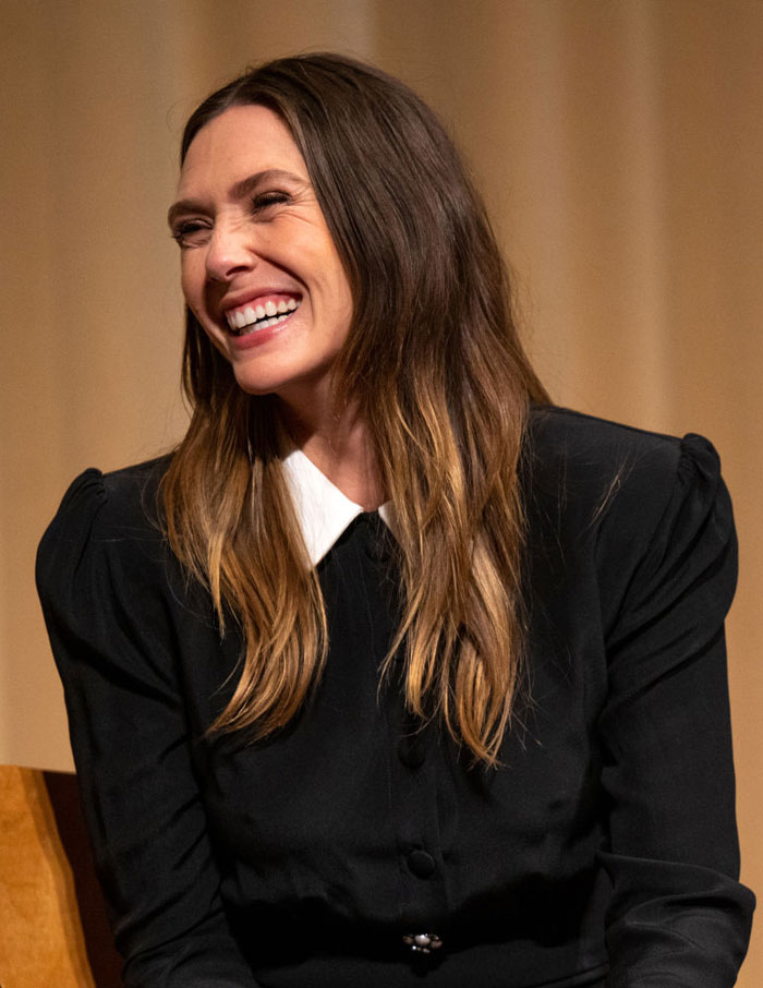  A B list celebrity, Elizabeth Olsen, is smiling and laughing while seated at an event, wearing a black dress with a white collar and long flowing hair.