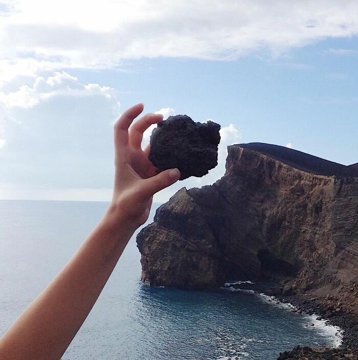 Hand holding a rock that mimics a cliff, creating a surreal perspective against the ocean backdrop.