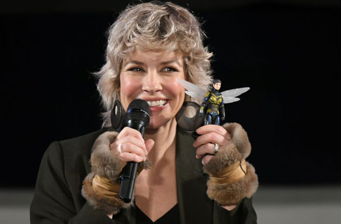 A B list celebrity, Evangeline Lilly, is smiling while holding a small action figure of her character during a public event, wearing a black jacket with fur-trimmed sleeves and speaking into a microphone.