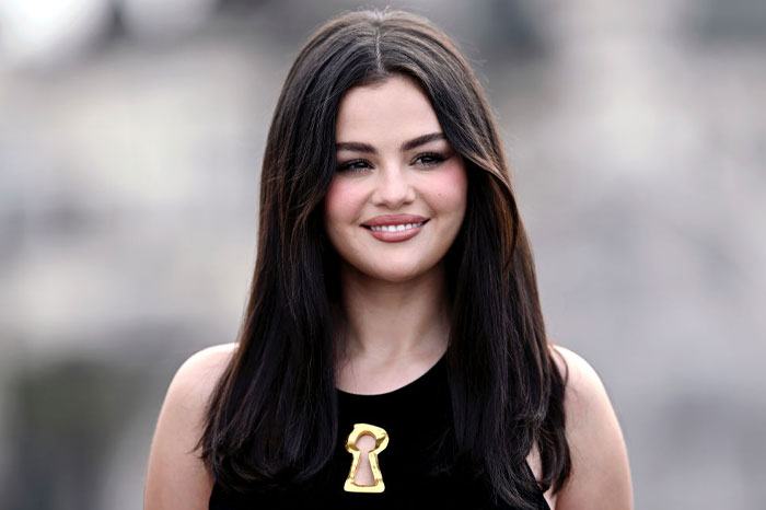 Selena Gomez, wearing a black dress with a gold keyhole emblem, smiles softly while standing outdoors with her long dark hair down.