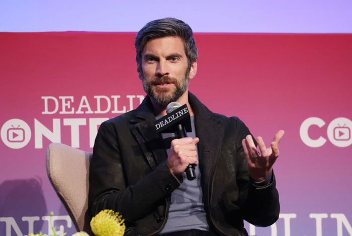  A B list celebrity, Wes Bentley, is speaking at a Deadline event, holding a microphone and gesturing with his hand while sitting against a branded backdrop.