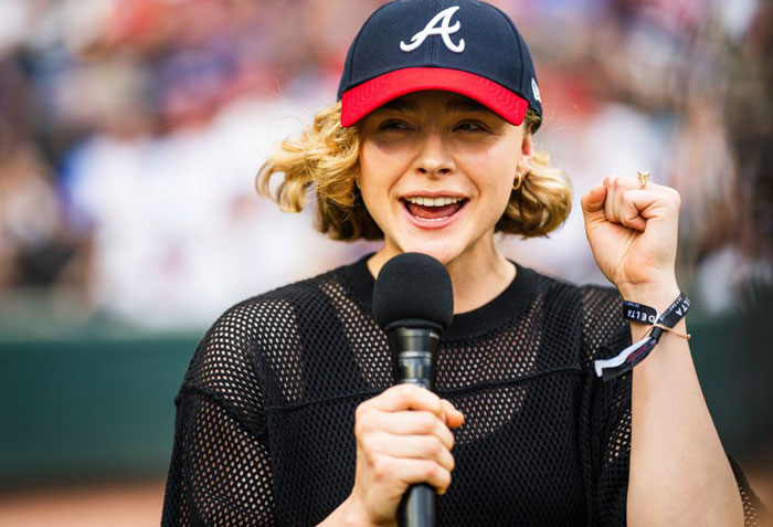  A B list celebrity, Chloë Grace Moretz, is speaking into a microphone while wearing a black mesh shirt and an Atlanta Braves baseball cap at a sports event, raising her fist enthusiastically.