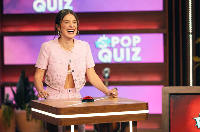  A B list celebrity, Millie Bobby Brown, is laughing while participating in a game show. She is wearing a pink cropped outfit and standing behind a quiz podium, with the words "Pop Quiz" visible on a screen in the background.