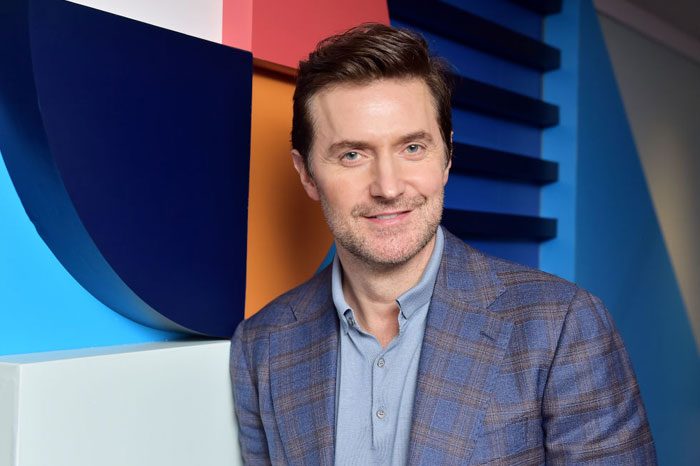  A B list celebrity, Richard Armitage, is smiling while leaning against a colorful geometric wall, wearing a blue plaid blazer and light blue shirt during an indoor event.