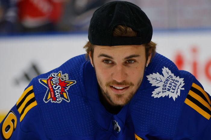 Justin Bieber, wearing a blue Toronto Maple Leafs jersey and a backwards black cap, leans forward and smiles during a hockey event.