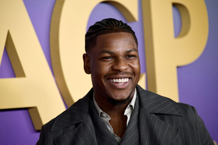 A B list celebrity, John Boyega, is smiling at an event, wearing a gray pinstripe suit and a white shirt, standing in front of a colorful NAACP backdrop.