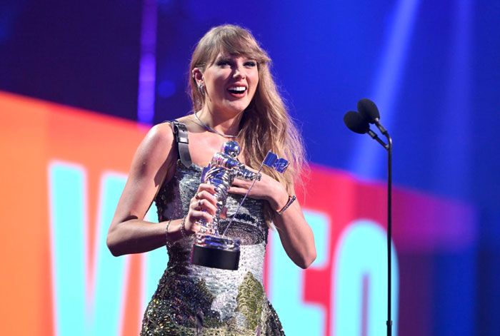 Taylor Swift, wearing a glittering dress, smiles while holding a silver award statue on stage during an award ceremony with a microphone nearby.