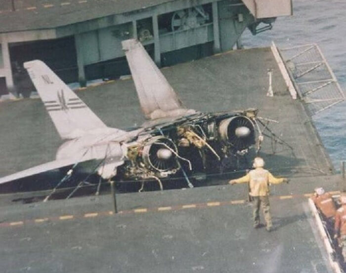 Damaged aircraft on carrier deck with crew member nearby; incident looks expensive due to visible destruction.