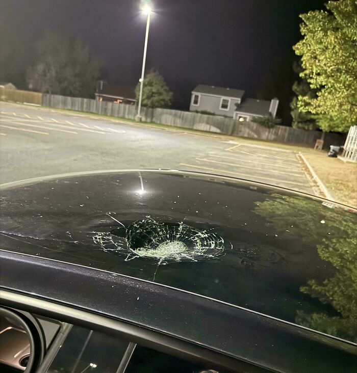 Car roof with a large, circular crack in the glass parked under a streetlight at night.