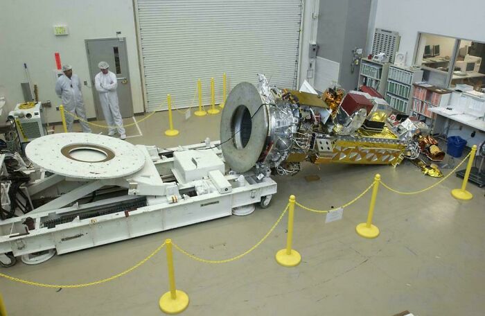 A damaged satellite lies on the floor in a lab, surrounded by yellow barriers, possibly an expensive incident.