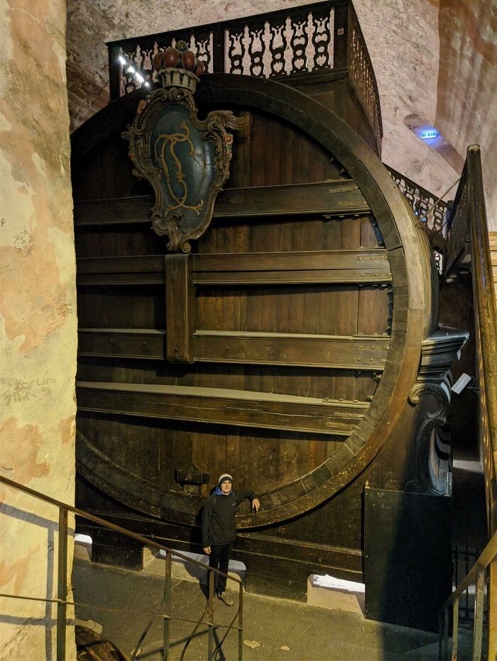 A person standing next to an absolute unit of a massive wine barrel inside a historic cellar.