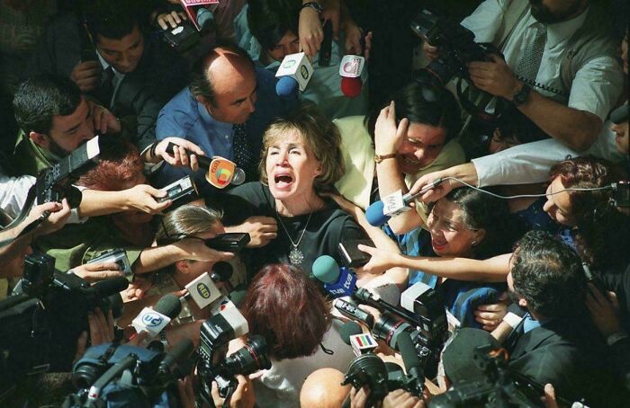 A woman surrounded by journalists with microphones, evoking a scene from a Renaissance painting.