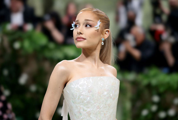 Ariana Grande, wearing an elegant white gown with feathered eye accents, looks to the side at a formal event with a blurred green background.