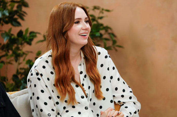 A B list celebrity, Karen Gillan, is smiling while seated, wearing a white polka dot blouse during an indoor event, with plants visible in the background.