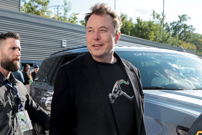 Elon Musk, wearing a black jacket and a graphic t-shirt, walks outdoors next to a vehicle with security personnel nearby, smiling slightly.