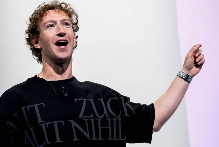 Mark Zuckerberg, wearing a black shirt with large text and a watch, gestures while speaking on stage during a presentation, smiling slightly.