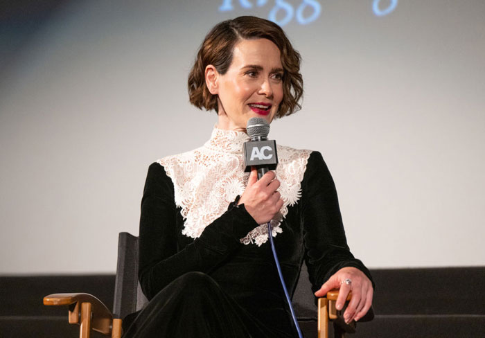 A B list celebrity, Sarah Paulson, is speaking into a microphone while seated at an event. She is wearing a black dress with a white lace collar.