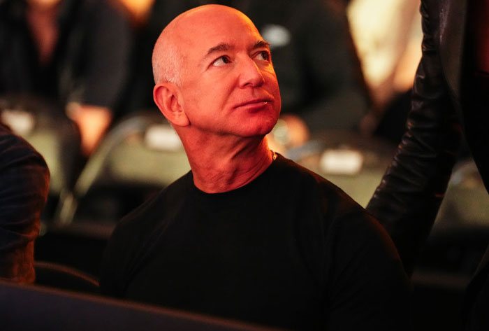 Jeff Bezos, wearing a black shirt, looks to the side while seated at an event, with a focused expression and dim lighting in the background.